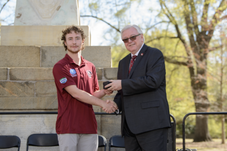 A young man with curly hair in a maroon polo shirt shakes hands with an older man in a suit, who is handing over a small box, in front of a monument with trees in the background.